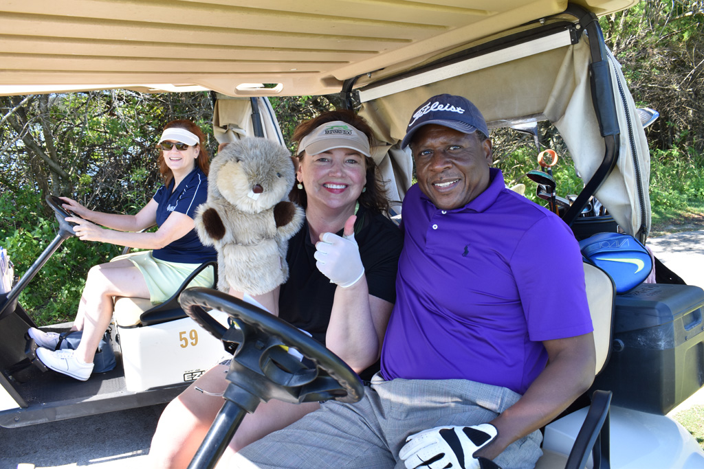 Three golfers sitting in two different gold carts smiling at the camera. One lady has a gopher golf cover on her hand that she is displaying to the camera.