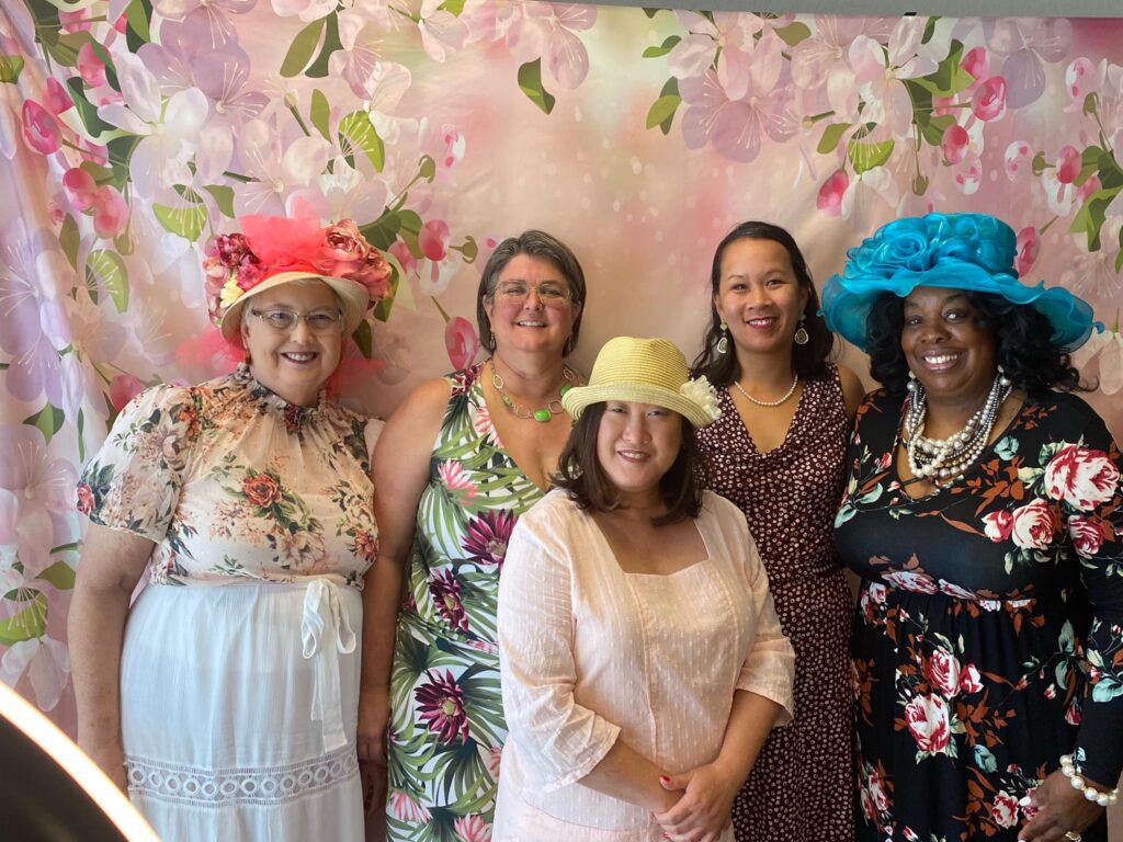 Five women stand together for a photo at the High Tea Fundraiser. Three of the women are in festive hats. These women stand behind a pink floral background.