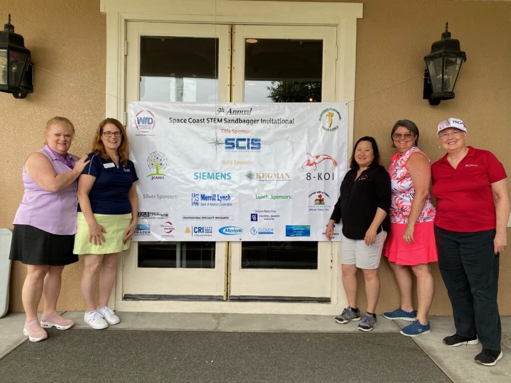 Five women stand by the Space Coast STEM Sandbagger Invitational banner with all of the listed sponsors.