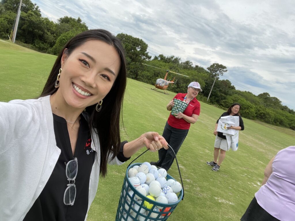 A women is taking a selfie with a bucket of gold balls in her hand. There are two other people in the background. A lady in a red shirt holds a bucket of golf balls and the lady behind her holds a sponsorship banner.